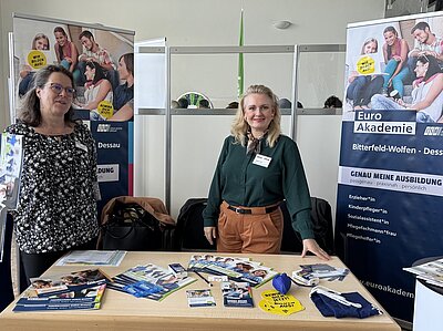 Zwei Mitarbeiterinnen der Euro Akademien Bitterfeld-Wolfen und Dessau präsentieren ihre Schulen am Messestand. Afu dem Tisch liegen verschiedene Infobroschüren aus, hinter dem Stand sind die Roll-ups mit den angebotenen Ausbildung aufgestellt.