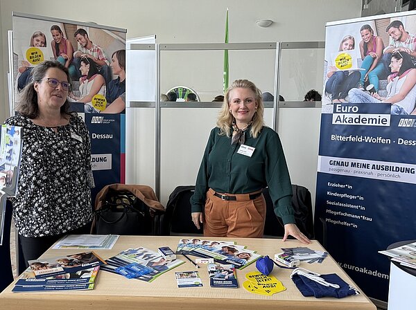 Volles Haus bei der Bildungsmesse in Bitterfeld-Wolfen Zwei Mitarbeiterinnen der Euro Akademien Bitterfeld-Wolfen und Dessau präsentieren ihre Schulen am Messestand. Afu dem Tisch liegen verschiedene Infobroschüren aus, hinter dem Stand sind die Roll-ups mit den angebotenen Ausbildung aufgestellt.