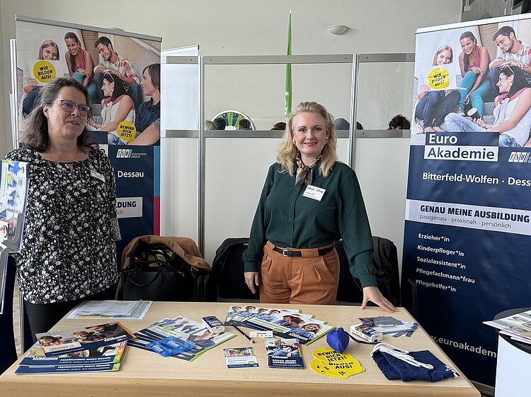 Volles Haus bei der Bildungsmesse in Bitterfeld-Wolfen Zwei Mitarbeiterinnen der Euro Akademien Bitterfeld-Wolfen und Dessau präsentieren ihre Schulen am Messestand. Afu dem Tisch liegen verschiedene Infobroschüren aus, hinter dem Stand sind die Roll-ups mit den angebotenen Ausbildung aufgestellt.