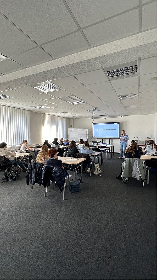 SCHAU REIN! – Woche der offenen Unternehmen Sachsen Eine Frau steht vorne im Klassenraum vor einer Präsentationsfolie mit dem Titel „Staatlich anerkannte/r Erzieher/in“, während die Teilnehmenden an Tischen sitzen und zuhören.