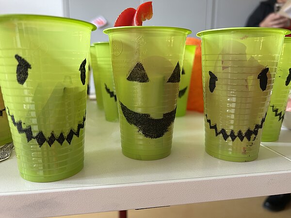 Neongrüne Plastikbecher mit aufgemalten Gruselgesichtern; auf dem Tisch arrangiert, teils mit Obstdeko.