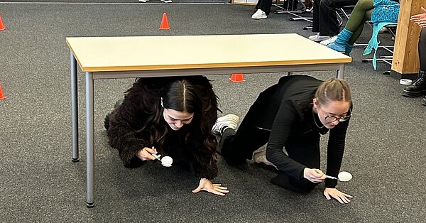 Zwei Schülerinnen krabbeln mit einem Ei auf einem Löffel unter einem Tisch durch.