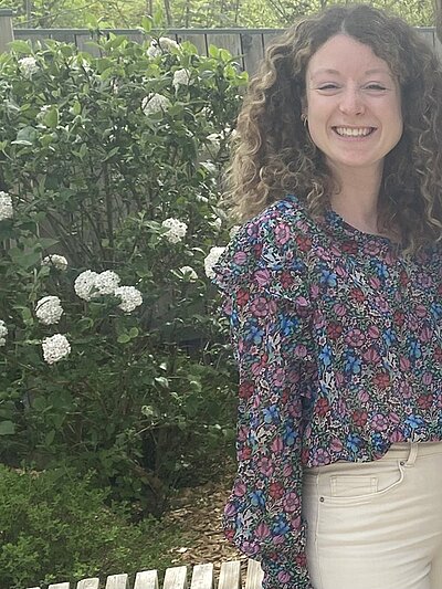 Absolventin der Euro Akademie Würzburg, Paula Krug, trägt eine helle Hose, eine Bluse mit floralem Muster, ihre lockigen Haare offen, hinter ihr eine Hecke und weiße Hortensien, sie lächelt in die Kamera für ein Foto.
