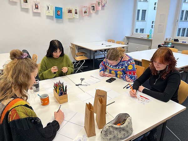 Vier Schülerinnen, von denen eine einen Weihnachtspulli trägt, sitzen an einem großen Tisch und malen Comics. Auf dem Tisch liegen Stifte, ein Handy, eine Schere, Tassen und Tüten. Eine Türe ist mit "Protagonist" beschriftet.