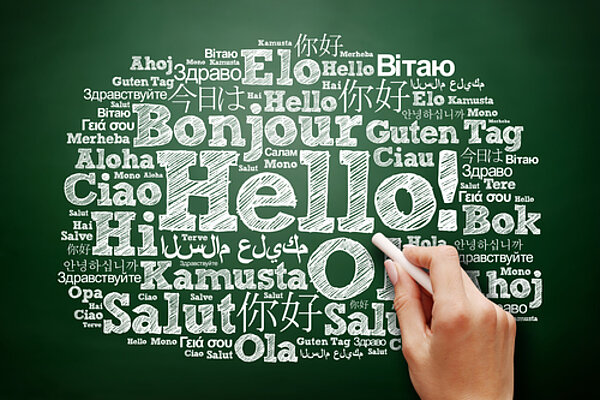In einer Grafik ist wie mit weißer Kreide auf einer grünen Tafel das Wort "Hallo" in verschiedenen Sprachen kreisförmig angeordnet, wobei "Hello!" in der Mitte am größten geschrieben ist und daher hervorsticht. Eine Hand mit einem Kreidestück scheint etwas dazu zu schreiben.
