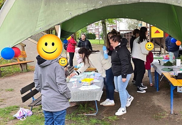 Unter einem großen, grünen Pavillon stehen mehrere Personen an einem Tisch mit gemustertem Tischtuch. Auf dem Tisch liegen Bastelmaterialien und bunte Stifte. Im Hintergrund sind weitere Personen, ein Spielplatz und bunte Luftballons zu sehen. Die Szene spielt im Freien auf einer Wiese.
