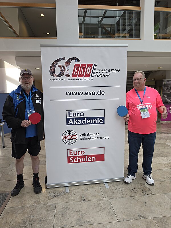 Zwei Männer mit Tischtennisschlägern stehen links und rechts neben einem Banner der ESO Education Group in einem Innenbereich.