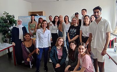 Eine Gruppe Schülerinne und Schüler hat sich in einem Klassenzimmer zu einem Gruppenbild aufgestellt. Im Hintergrund steht eine Staffelei mit einem Bild darauf, daneben noch eine Zimmerpflanze.
