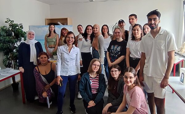 Unterrichtsprojekt “Feria de empresas” Eine Gruppe Schülerinne und Schüler hat sich in einem Klassenzimmer zu einem Gruppenbild aufgestellt. Im Hintergrund steht eine Staffelei mit einem Bild darauf, daneben noch eine Zimmerpflanze.