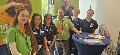 Sechs Mitarbeitende und Auszubildende der Euro Akademie stehen lächelnd an einem Messestand hinter einem blauen Stehtisch mit Flyern, Kugelschreibern und Schlüsselbändern vor farbigen Roll-ups.