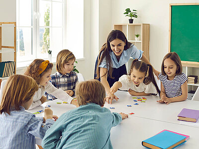 Eine junge Lehrerin mit langen, braunen Haaren, die einen dunkelblauen Rock und eine hellblaue Bluse trägt, beugt sich über einen Tisch, an dem sechs Schülerinnen und Schüler im Grundschulalter sitzen. Diese greifen nach bunten Buchstaben, die auf dem Tisch liegen. Im Hintergrund sind Regale mit Büchern und Pflanzen sowie ein Tafel erkennbar.