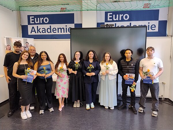 Eine Reihe lächelnder Menschen in festlichen Kleidern steht vor einem großen Bildschirm. Die Menschen halten Blumen und Zeugnisse. An der Wand dahinter ist in dunkelblau zweimal das Logo der Euro Akademie zu sehen.