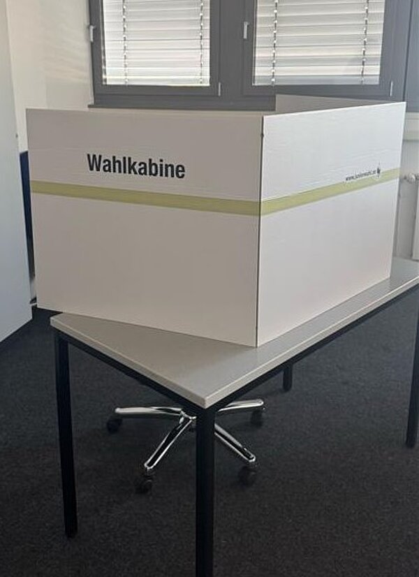 Testwahlen an der Euro Akademie Aschaffenburg am 19. Februar 2025 Eine weiße Wahlkabine mit der Aufschrift „Wahlkabine“ steht auf einem Tisch in einem Wahlraum. Sie bietet Wählern eine private Möglichkeit zur Stimmabgabe.
