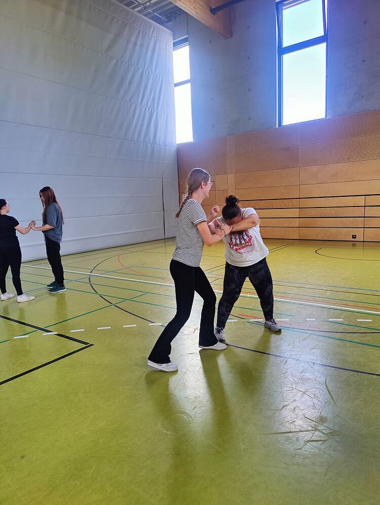 Zwei Frauen rangeln miteinander in einer Sporthalle mit gelbem Boden und Holzpaneelen an den Wänden. Die eine Frau trägt ein gestreiftes T-Shirt und eine schwarze Sporthose. Die andere Frau trägt ein helles T-Shirt mit Aufdruck und dunkle Leggins. Im Hintergrund sind zwei weitere Personen zu sehen, die ebenfalls Übungen zur Selbstverteidigung durchführen.