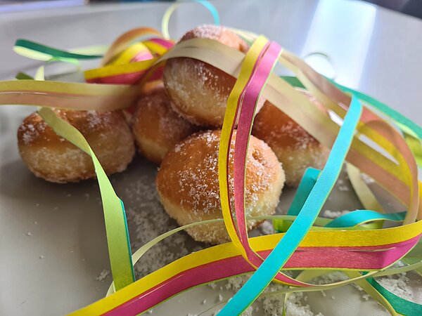 Helau und Alaaf in Rochlitz Ein Teller mit kleinen, goldbraunen Berlinern (Krapfen), bestreut mit Puderzucker, liegt auf einem Tisch. Bunte Papier-Luftschlangen sind dekorativ über das Gebäck gelegt und verstärken die festliche Faschingsstimmung.