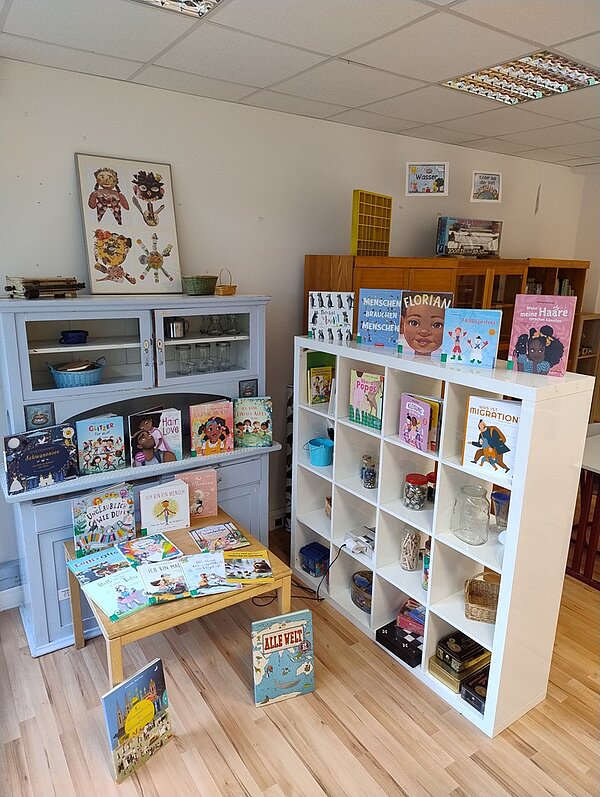 Studierende der Fachschule gestalten eindrucksvolle Ausstellung Auf einem weißen Regal sind zahlreiche Bücher einsortiert. Auf der Retro-Anrichte in der Farbe taubengrau stehen ebenfalls Kinderbücher sowie auf einem kleinen, weißen Tisch.