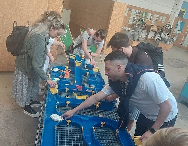 Ausflug ins Wasserzentrum Bitterfeld-Wolfen Auf einem Tisch mit Gittern ist eine Art Parcours aus blauem Plastik aufgebaut. In den Halbrohren fließt Wasser. Fünf junge Menschen mit Rucksäcken schieben kleine Plastikboote durch den Parcours. Im Hintergrund sind im selben Raum noch andere Stationen aufgebaut, an denen aber niemand steht.