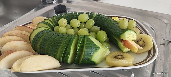 Gesund und kreativ Auf einem Teller ist eine Salatgurke rund platziert und als Schlange, mit Obstzunge gestaltet. Zentriert liegen grüne Trauben, am Salatkopf liegen Kiwischeiben und rundherum sind Apfelschnitze platziert.