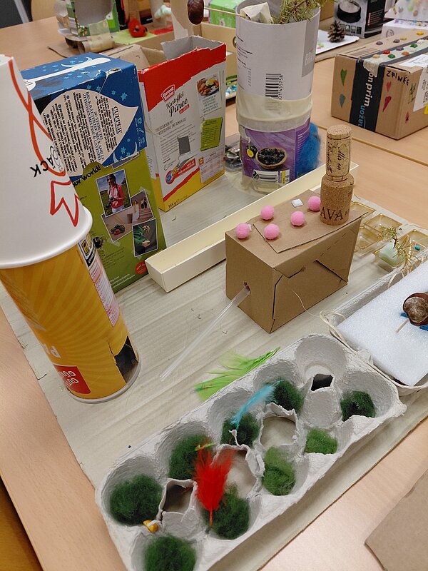 Seitliche Ansicht derselben Bastelarbeit: Man sieht mehrere aufgeklebte Verpackungskartons, eine gelbe Chipsdose, den Kartonturm mit Korken und Pompons sowie den Eierkarton mit grüner Wolle und Federn, alles arrangiert auf einer großen Pappe.