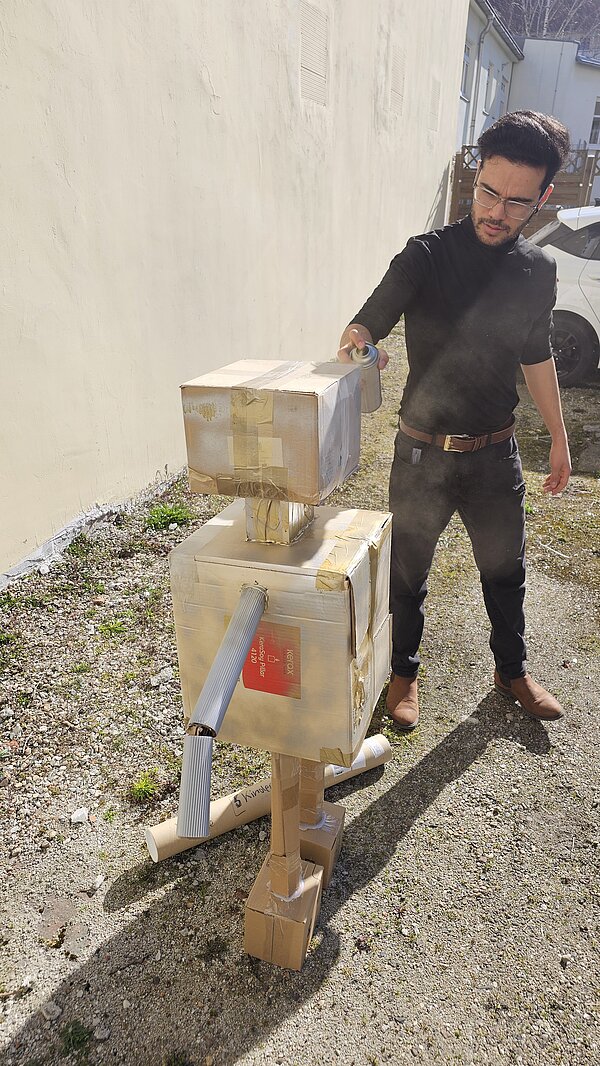 Robotik: Innovative Ansätze in der Pflege Ein Mann in dunkler Kleidung besprüht einen Roboter aus verschiedenen Kartons mit goldener Farbe.
