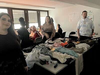 Mehrere Tische sind mit einer Vielzahl von Kleidungsstücken in unterschiedlichen Farben und Materialien bedeckt, darunter Pullover, T-Shirts und Hosen. Im Vordergrund stehen die Auszubildenden, die Kleidung begutachten und untereinander tauschen. Im Hintergrund sitzen weitere Menschen an einem Tisch und führen Gespräche. Die Veranstaltung findet in einem hellen Raum mit großen Fenstern und Jalousien statt. An der Wand hängt ein Plakat mit handgeschriebenem Text. Die Szene zeigt einen nachhaltigen Kleidertauschmarkt, organisiert von den Auszubildenden der Euro Akademie Wiesbaden, der zum umweltbewussten Konsum und sozialen Austausch beiträgt.