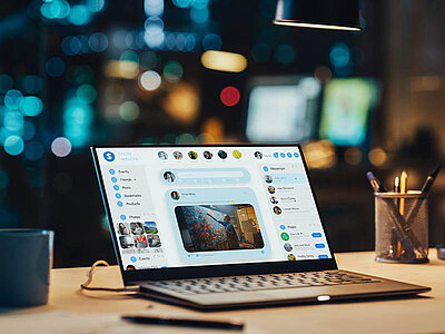 Ein aufgeklappter Laptop steht auf einem Schreibtisch, auf dem auch eine Tasse und ein Glas mit Stiften stehen. Auf dem Bildschirm ist eine Webseite zu sehen. Im Hintergrund befinden sich mehrere Bildschirme, die unscharf dargestellt werden.