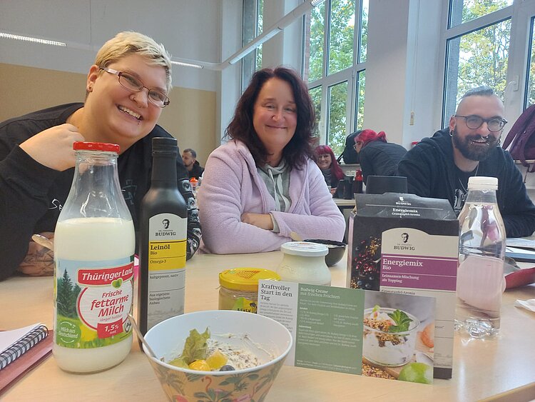 An einem Holztisch, auf dem eine Glasflasche mit Milch und ein Müsli steht, sitzen links zwei Frauen und rechts ein Mann, die lächeln.