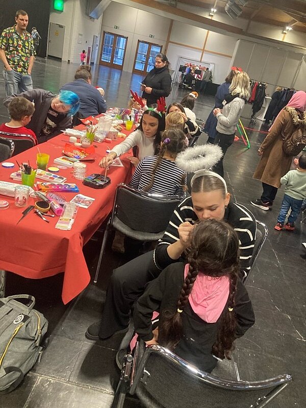 Eine Gruppe von Kindern und Erwachsenen sitzen an einem langen Tisch. Die Erwachsenen schminken die Kinder. Im Hintergrund sind weitere Erwachsene und weitere Kinder zu sehen, die sich in einem Gemeinschaftsraum aufhalten. Der Tisch ist mit Schminkutensilien bedeckt.