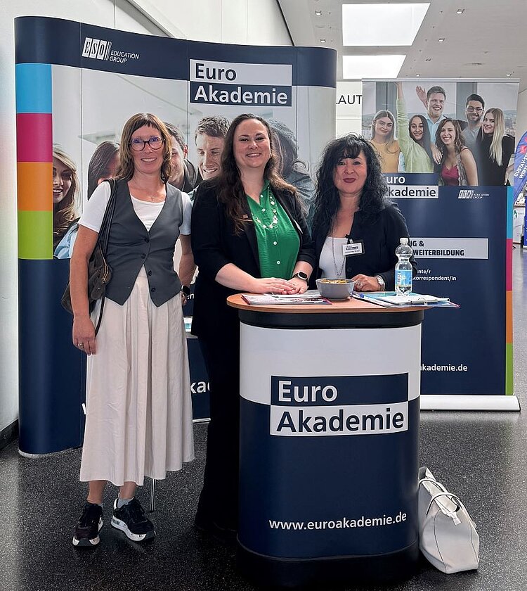 Drei Frauen stehen lächelnd vor einem Messestand der Euro Akademie. Der Stand ist mit Roll-ups und einem runden Tisch mit der Aufschrift 'Euro Akademie' und der Website www.euroakademie.de gestaltet.