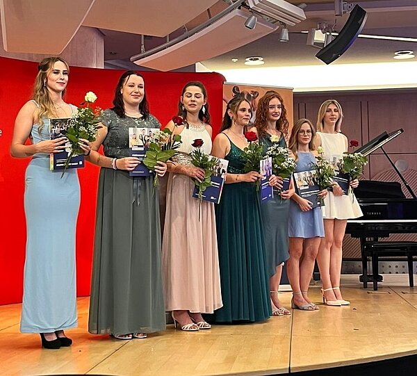 Feierlicher Abschied: Exmatrikulation der Absolvent*innen 2025 Eine Gruppe von Personen steht auf einer Bühne und hält Blumen und Zertifikate. Sie sind elegant gekleidet und lächeln. Im Hintergrund ist ein rotes Banner und ein Klavier zu sehen.