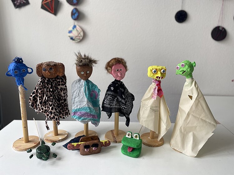 Auf einem Tisch stehen verschiedene Handpuppen auf Holzständern. Diese haben grüne, gelbe, rote, schwarze und blaue Köpfe und jeweils Körper aus Stofffetzen, darunter einen mit Leopardenmuster.