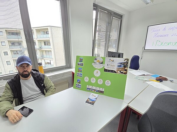 In einem hellen Raum mit großen Fenstern sitzt ein Auszubildender an einem Tisch. Auf dem Tisch steht ein grünes Plakat mit mehreren Fotos, Logos und Textfeldern, die in verschiedenen Anordnungen aufgeklebt sind. Im Hintergrund befinden sich weitere Tische, Stühle und ein Bildschirm, auf dem handschriftlich Text steht. Draußen sind Wohngebäude zu sehen.