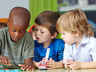Drei Jungen unterschiedlicher ethnischer Herkunft sitzen nebeneinander an einem Tisch und bauen gemeinsam mit LEGO-Steinen. Sie sind konzentriert und arbeiten zusammen an einem Projekt.