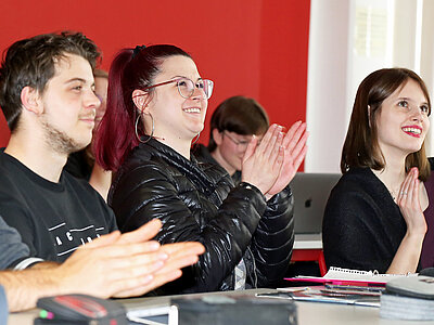 Mehrere Studierende sitzen im Unterricht vor roter Wand und klatschen lächelnd, auf dem Tisch liegen Hefte, Stifte und Federmappen.