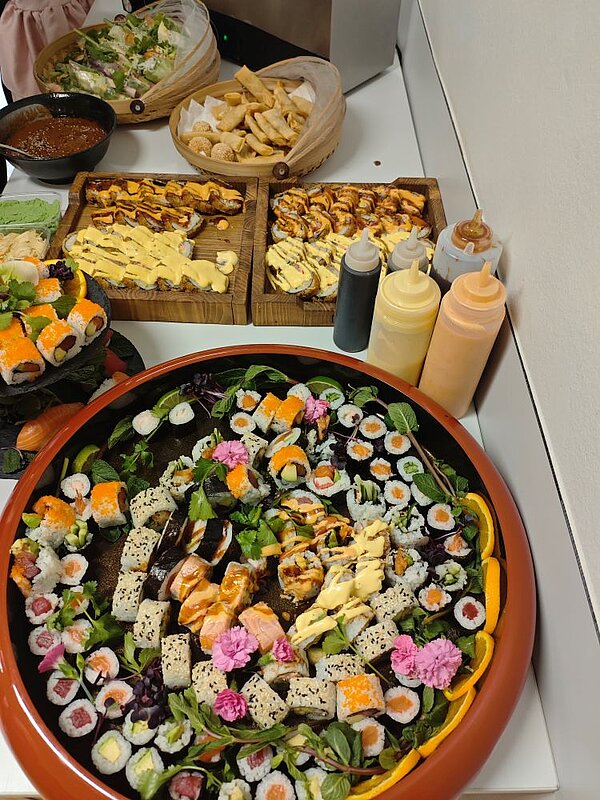 Ein reichhaltiges Buffet mit einer Vielzahl asiatischer Gerichte: Sushi-Rollen, Sashimi, Tempura, Frühlingsrollen und anderen kulinarischen Köstlichkeiten. Die Speisen sind auf großen Platten und in Schalen angerichtet, umgeben von Saucenflaschen und garniert mit Blättern und essbaren Blüten.