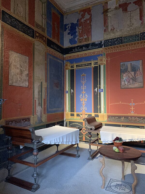 Eintauchen in die römische Kultur In der Ecke eines Raumes im Pompejanum stehen zwei Liegen mit Tücher darüber, daneben ein kleine Holztisch. Die Wände sind bunt bemalt, auch auf dem Boden befinden sich bemalte Fliesen.