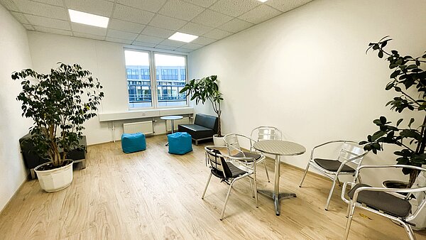 Gemütlicher Ruheraum mit zwei großen Zimmerpflanzen, kleinen runden Metalltischen, Stühlen, zwei Sitzbänken und türkisfarbenen Sitzsäcken vor einem Fenster.