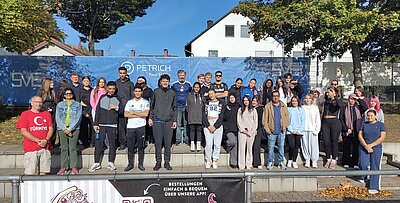 Eine große Gruppe von Menschen steht auf einer Treppe vor einem blauen Banner mit der Aufschrift 'PETRICH Real Estate'. Die Gruppe posiert für ein Foto im Freien und die Sonne scheint.