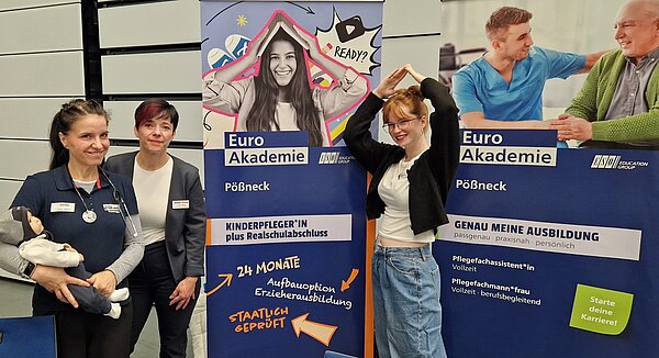 Drei Personen stehen vor Roll-ups der Euro Akademie; eine Person formt mit den Armen ein Dach über dem Kopf passend zum Motiv auf dem Banner.