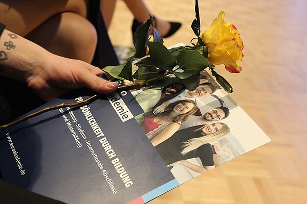 Sicht auf eine Hand, die eine Euro Akademie-Broschüre sowie eine gelbe Rose in den Händen hält.