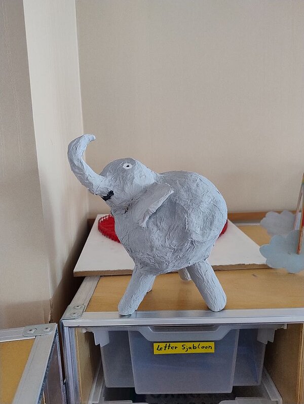 Ein selbstgebastelter Elefant aus grauer Papiermache, der auf einem Regalbrett in einem Klassenzimmer oder einer Werkstatt steht. Im Hintergrund sind Aufbewahrungsboxen und ein Schrank zu sehen.