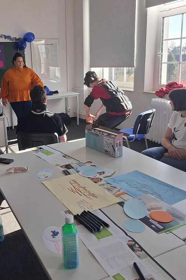 Gruppe von Menschen in einem hellen Seminarraum: Eine Person versucht gerade vom Stuhl aufzustehen, während sie Kopfhörer, eine Schutzbrille und eine schwarze Weste trägt. Auf dem Tisch liegen verschiedene Materialien wie Flyer, Stifte und farbige Pappkreise. Im Hintergrund sitzen weitere Personen auf Stühlen, während eine Person im orangefarbenen Oberteil vor einer Tafel steht und etwas zu erklären scheint.