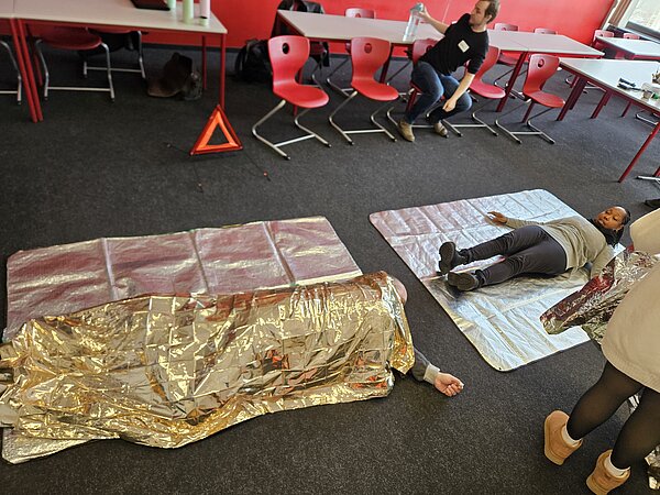 Praxisübung: Teilnehmende liegen bzw. sitzen auf Isomatten und werden mit Rettungsdecken versorgt; Warndreieck und Materialien im Hintergrund.