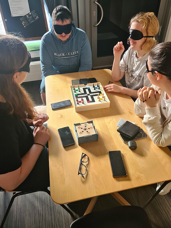 Vier Schüler*innen mit Augenmaske sitzen an einem Holztisch und spielen blind Mensch-Ärgere-Dich-Nicht.
