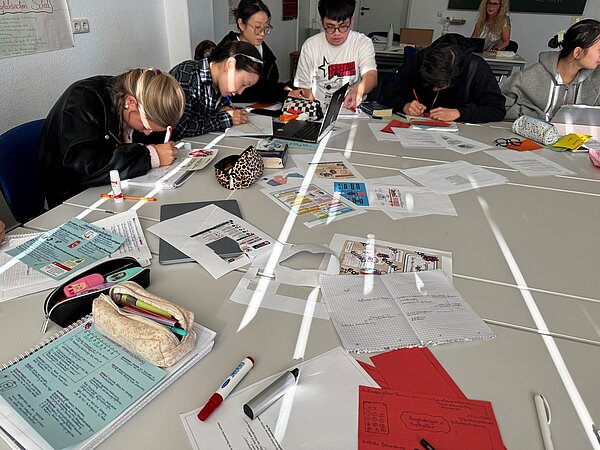 Mehrere Schülerinnen und Schüler sitzen gemeinsam an einem großen Tisch und bearbeiten Unterrichtsmaterialien. Auf dem Tisch liegen Notizen, Arbeitsblätter, Stifte, Tablets und selbst gestaltete Lernkarten.