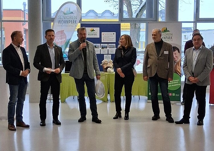 Sechs Personen stehen in einer Reihe in einem Innenraum. Im Hintergrund befinden sich ein grüner Tisch mit Flyern und Broschüren, mehrere Aufsteller mit der Aufschrift 'SENIOREN WOHNPARK', sowie weitere Informationsmaterialien. Die Personen tragen überwiegend Anzüge oder Blazer und halten teilweise Mikrofone oder Papiere in den Händen. Große Fenster lassen Tageslicht herein, und draußen sind Gebäude und ein Baum zu sehen.