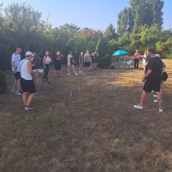 Sportfest – mal anders Auf einer Wiese stehen Plastikflaschen in einer Reihe, außen herum einige Schüler*innen, die einen älteren Person zuhören, die offenbar gerade die Regeln erklärt. Im Hintergrund steht ein Tisch und daneben ein türkisfarbener Sonnenschirm