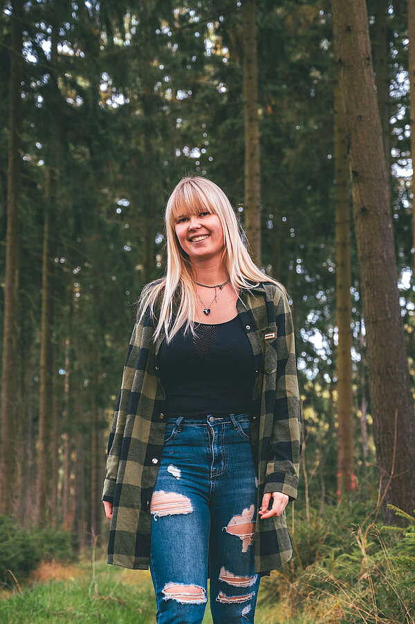 eine junge Frau mit blonden Haaren steht vor einem Wald und lächelt in die Kamera.