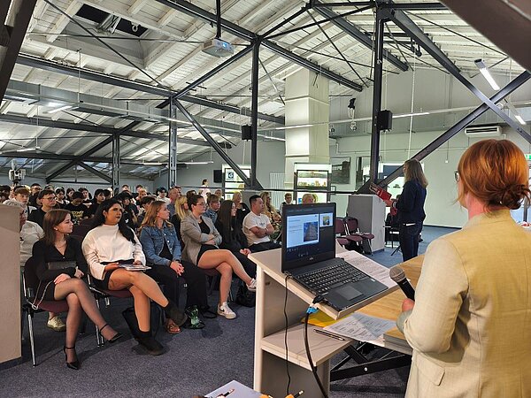 Herzlicher Abschied, freudiger Anfang: Pflege-Absolvent*innen geehrt und neue Auszubildende willkommen geheißen Eine Frau in einem hellen Blazer steht an einem Podium mit Laptop vor einem Publikum und hält ein Mikrofon in der Hand. Eine andere Frau spricht zu einer Gruppe von Zuhörern, die auf Stühlen in einer großen Halle sitzen..