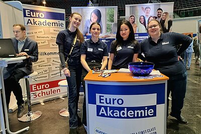Das Bild zeigt den Messestand der Euro Akademie und der Euro-Schulen. Im Vordergrund steht das Pult mit Logo und dahinter sind vier Auszubildender im T-Shirt der Euro Akademie zu sehen, die freundlich in die Kamera blicken.